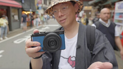 Photographer holding a blue compact digital camera while standing in an urban street.