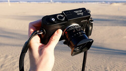 Hand holding a rangefinder camera with attached lens against a beach background.