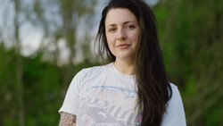 Woman with long dark hair wearing a white graphic t-shirt, photographed outdoors with soft natural light.