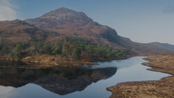 Mountain reflected in still lake water with forested slopes and moorland surrounding the shoreline.