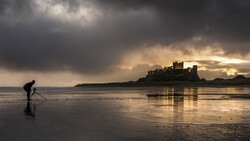 Photographer with tripod on wet beach at sunset, castle silhouetted against dramatic sky across the water.