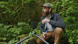 Cinematographer crouching beside a creek in dense forest vegetation, holding audio recording equipment.