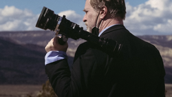 Photographer in dark jacket holding a telephoto lens to his eye, photographing a vast canyon landscape under blue sky.