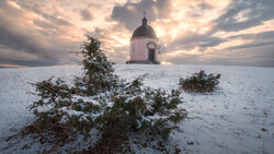 Snow-covered landscape with a domed chapel and bare shrubs under dramatic cloudy sky.
