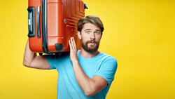 Bearded man in light blue t-shirt carrying red suitcase on shoulder against yellow background.