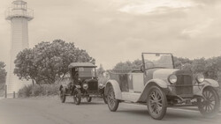 Two vintage automobiles parked near a lighthouse under overcast sky.