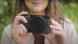 Woman holding a black Lumix compact camera outdoors with blurred green background.