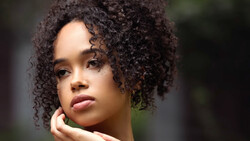 Portrait of a young woman with dark curly hair, looking toward the camera with a contemplative expression.