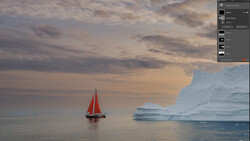 Red-sailed boat on calm water with large icebergs under a dramatic cloudy sky.