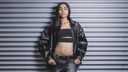 Woman in black leather jacket and crop top posing against corrugated metal wall.