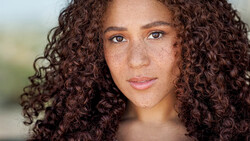 Portrait of a woman with long curly brown hair and freckles looking directly at camera.