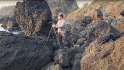 Photographer on tripod positioning among rocky coastal formations with water and forested hills in background.