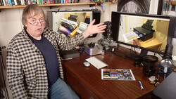 Photographer gestures toward camera equipment displayed on a computer monitor in a home studio setup.