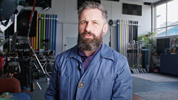 Bearded man in blue shirt standing in professional photography studio with lighting equipment and grip gear visible in background.
