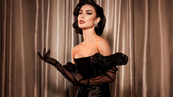 Woman in black off-shoulder velvet dress with sheer sleeves and long black gloves against draped neutral backdrop.