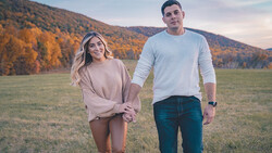 Couple walking hand-in-hand through an autumn field with forested mountains in the background.