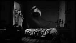 A figure suspended above a bed in a dramatically lit black and white interior scene.