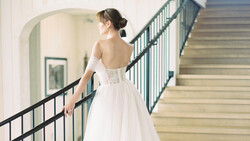 Bride in white off-shoulder gown descending staircase, hand on railing, viewed from behind.