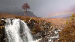 Waterfall cascades over rocky terrain with a solitary tree on the hillside and a rainbow arcing across the misty valley beyond.