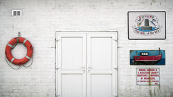White double doors on weathered wall with life preserver, festival sign, boat painting, and charity plaque.