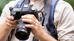 Photographer holding a DSLR camera with telephoto lens and blue camera strap.