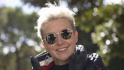 Person with white hair wearing round sunglasses and dark jacket with red and white striping, photographed outdoors with trees in soft focus background.