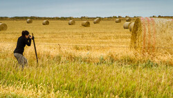 Photographer with camera on tripod capturing hay bales in rural field.