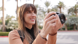 Woman holding a compact camera outdoors, smiling while composing a shot in a palm-tree lined park.