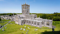 Medieval stone church with square tower and graveyard surrounded by green fields and trees under blue sky.