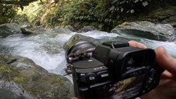 Photographer holding a mirrorless camera with wide-angle lens pointed at flowing stream surrounded by rocks and forest.