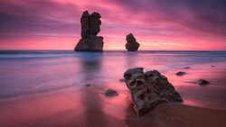Rocky sea stacks silhouetted against a vibrant pink and purple sunset reflected in calm shallow water.
