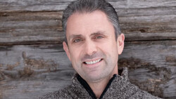 Man smiling at camera wearing gray knit sweater against weathered wood backdrop.