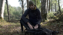 Man crouching beside backpack in forest clearing surrounded by tall trees.