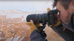 Photographer holding a mirrorless camera with telephoto lens on tripod, shooting snowy canyon landscape.