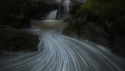 Long-exposure photograph of a flowing stream with silky water patterns curving around moss-covered rocks and a waterfall in the background.