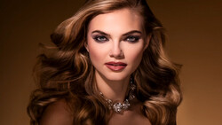 Woman with voluminous wavy brunette hair and dark red lipstick wearing a decorative statement necklace against a warm brown background.