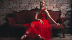 Woman in a red tulle gown seated on a burgundy velvet sofa against an exposed brick wall.