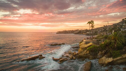 Rocky coastal cliff at sunset with palm trees and golden light reflecting on calm ocean waters.