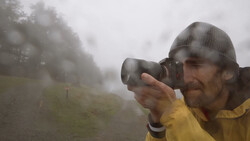 Photographer in yellow jacket and beanie shooting in heavy fog with binoculars-style camera positioning.
