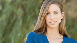 Woman with long brown hair wearing a blue top, photographed outdoors against blurred green foliage.