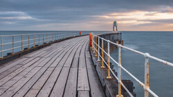 Wooden pier with white railings extending toward a lighthouse under dramatic cloudy sky at twilight.