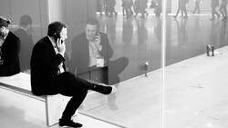 Businessman in dark suit sitting on white bench against glass window, speaking on phone while observing busy corridor reflection.