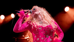 Performer in magenta sequined outfit and blonde curled wig singing into microphone on stage.
