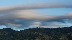 Dramatic lenticular clouds layered over a forested mountain landscape beneath blue sky.