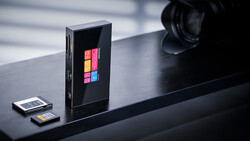 Vertical black device with colorful grid display sits on dark surface next to memory cards and camera lens.