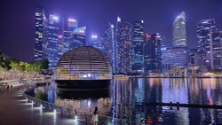 Modern dome structure on waterfront promenade with downtown city skyline reflected in calm water at night.