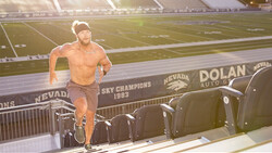 Shirtless runner in motion on a stadium press box with field and signage visible behind.