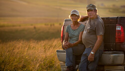 Two people in caps leaning against a truck bed in an expansive grain field during golden hour.