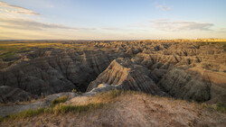 Badlands landscape with eroded buttes, deep ravines, and sparse prairie grass under golden hour light.