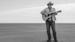 Man playing acoustic guitar while standing alone on a vast, flat desert plain under an overcast sky.